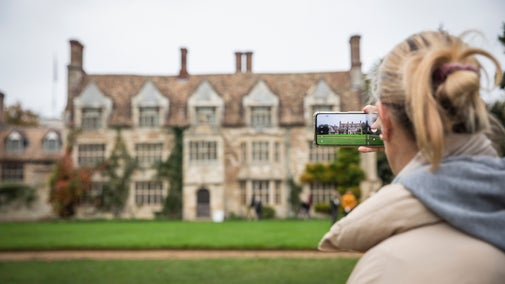 A lady takes a photo of the Anglesey Abbey house on her phone. The phone is in focus.